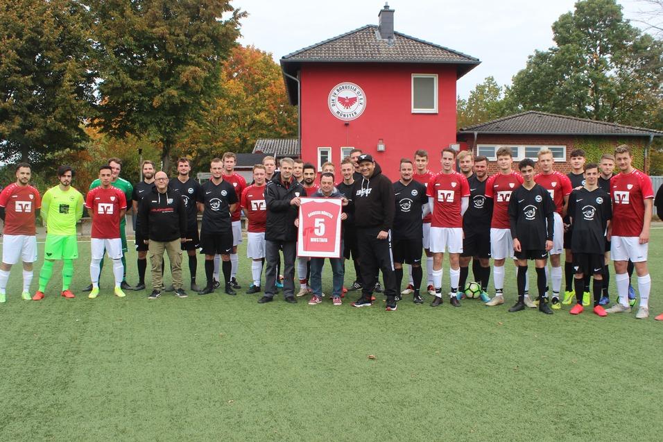 Gruppenfoto von zwei Fu&szlig;ballmannschaften in roten und schwarzen Trikots, stehend auf einem Kunstrasenplatz vor einem Vereinsgeb&auml;ude.