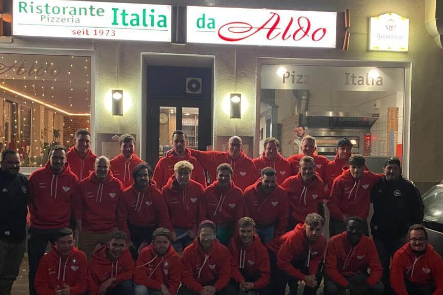 Gruppenfoto von 24 Personen in roten Sweatshirts vor dem Restaurant "da Aldo" mit beleuchteter Fassade.