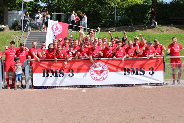 Gruppennfoto von Spielern in roten Trikots vor einem Banner mit der Aufschrift "BMS 3", alle jubeln in sonniger Umgebung.