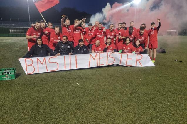 Gruppe von Spielern in roten Trikots posiert in der Nacht mit einem Banner &bdquo;BMS III Meister&ldquo; und Feuern im Hintergrund.