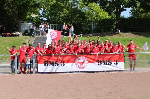 Gruppennfoto von Spielern in roten Trikots vor einem Banner mit der Aufschrift "BMS 3", alle jubeln in sonniger Umgebung.