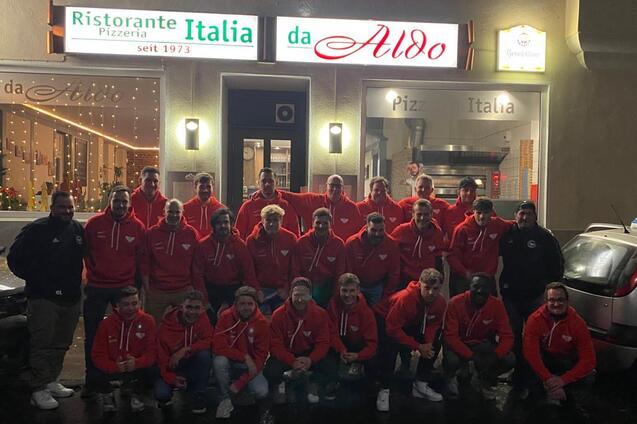Gruppenfoto von 24 Personen in roten Sweatshirts vor dem Restaurant "da Aldo" mit beleuchteter Fassade.