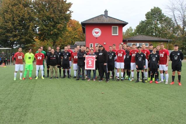 Gruppenfoto von zwei Fu&szlig;ballmannschaften in roten und schwarzen Trikots, stehend auf einem Kunstrasenplatz vor einem Vereinsgeb&auml;ude.