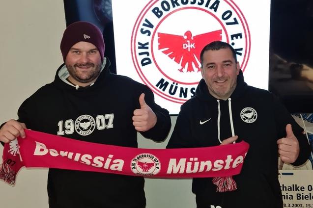 Zwei M&auml;nner zeigen Daumen hoch und halten einen Schal mit dem Schriftzug &bdquo;Borussia M&uuml;nster&ldquo; vor einem Vereinslogo.