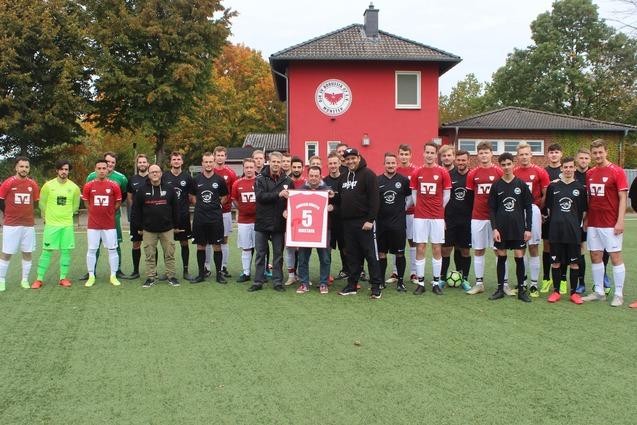 Gruppenfoto von zwei Fu&szlig;ballmannschaften in roten und schwarzen Trikots, stehend auf einem Kunstrasenplatz vor einem Vereinsgeb&auml;ude.