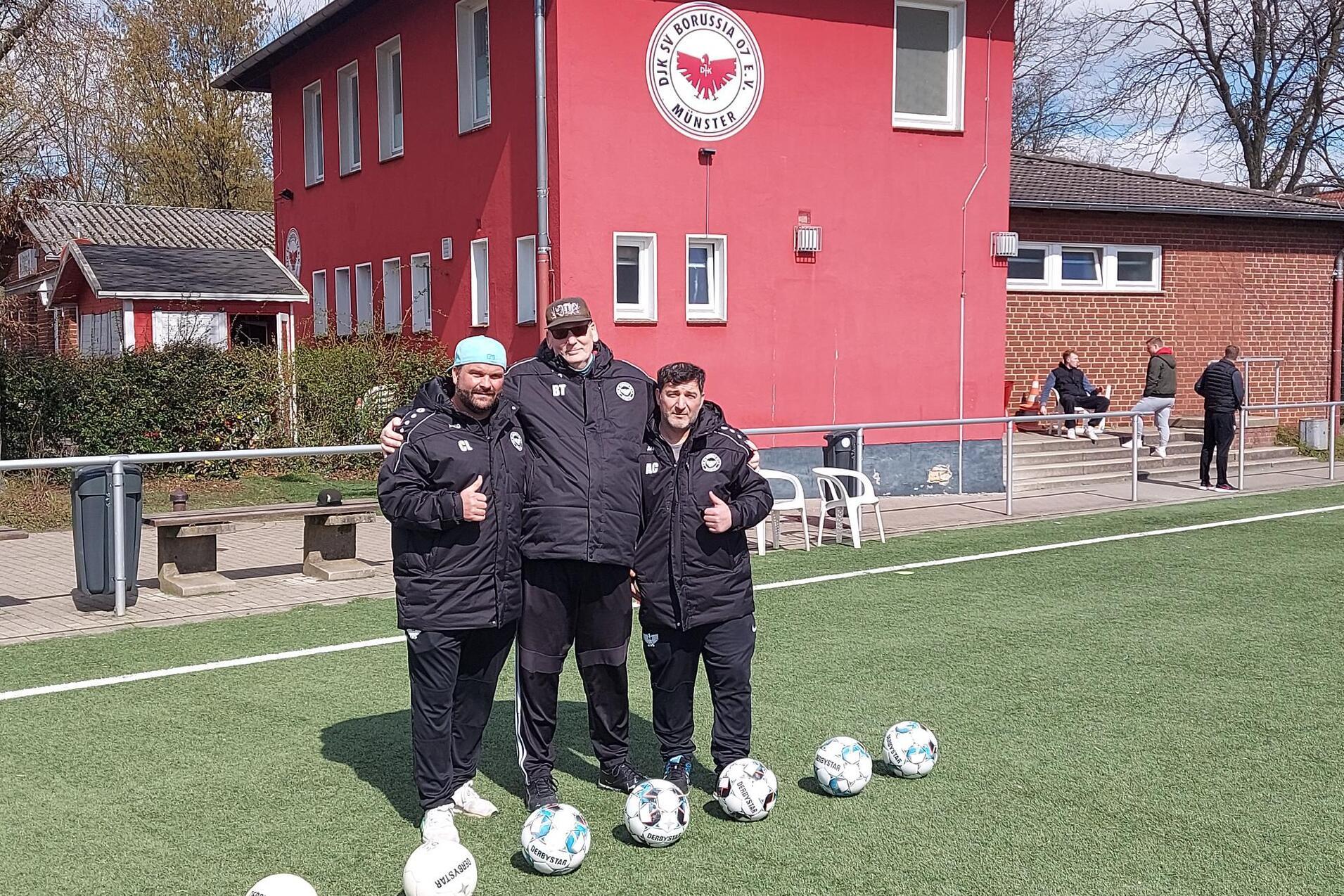 Drei Trainer in schwarzen Jacken stehen l&auml;chelnd auf einem Fu&szlig;ballfeld vor einem roten Vereinsgeb&auml;ude, Fu&szlig;ballsen liegen vor ihnen.