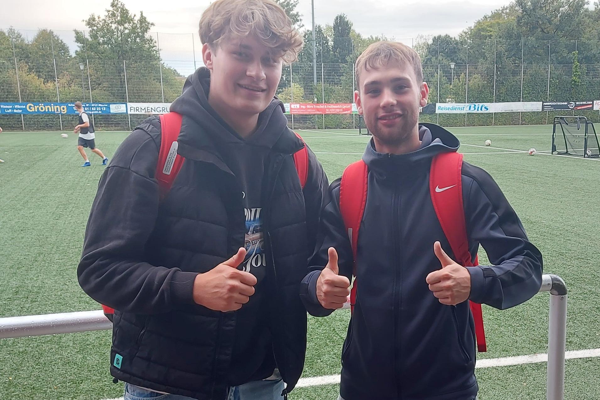 Zwei Jugendliche mit Rucks&auml;cken geben Daumen hoch, im Hintergrund ein Fu&szlig;ballfeld mit Spielern.