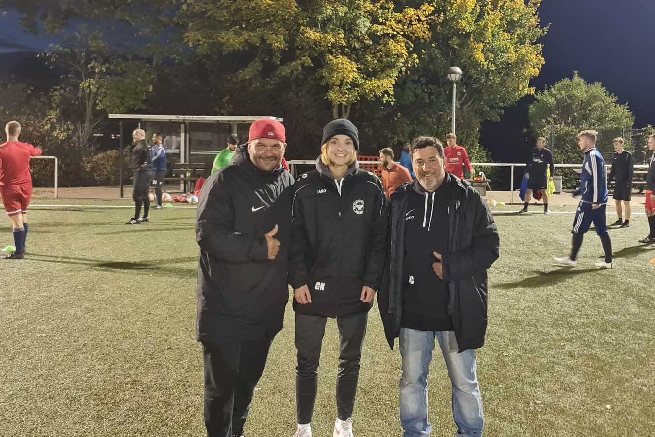 Drei Personen stehen auf einem Fu&szlig;ballplatz, umgeben von Spielern im Hintergrund unter Flutlicht.