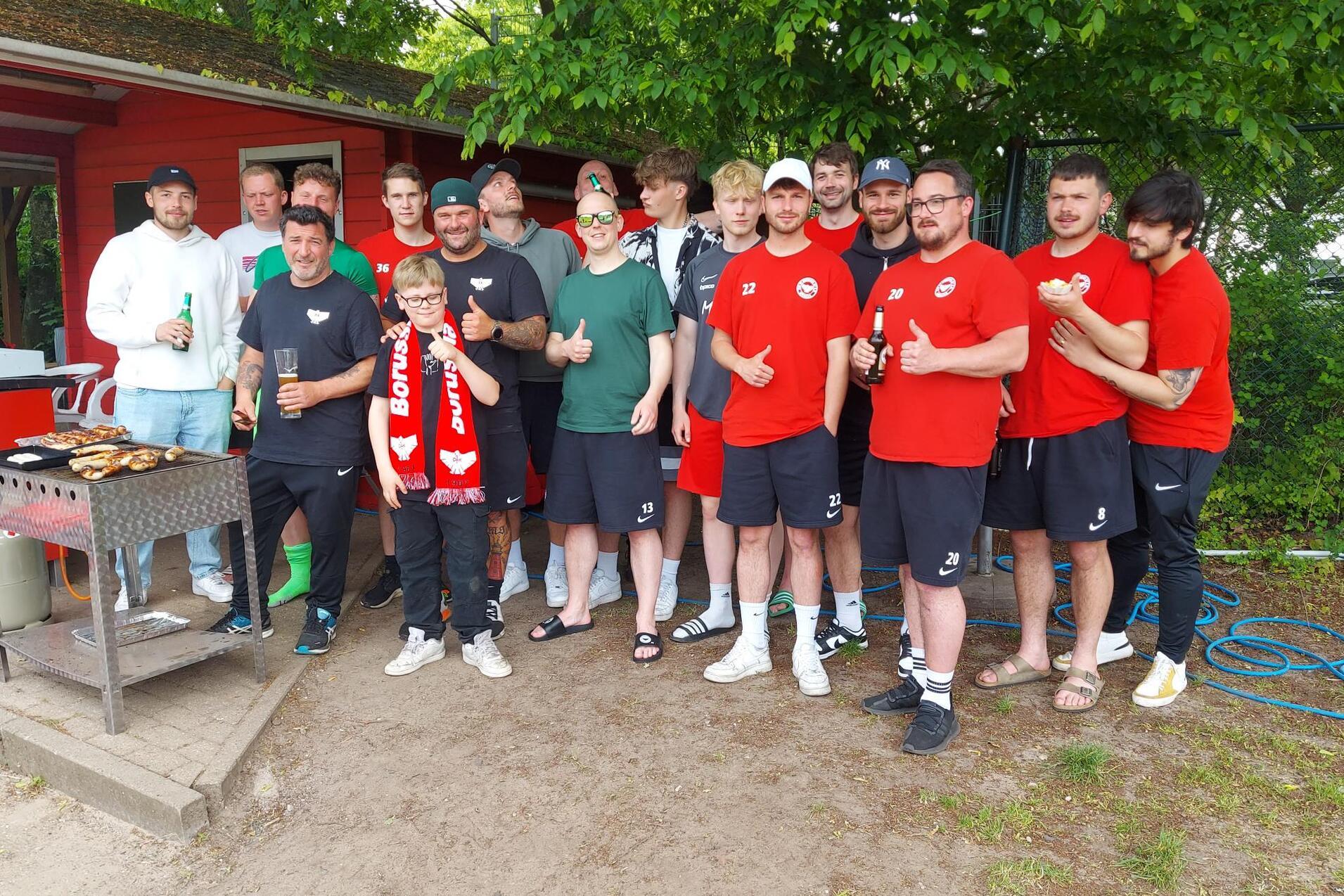 Gruppe von M&auml;nnern in roten T-Shirts versammelt um einen Grill, l&auml;chelnd und mit Getr&auml;nken in der Hand.