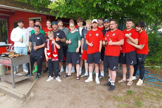 Gruppe von M&auml;nnern in roten T-Shirts versammelt um einen Grill, l&auml;chelnd und mit Getr&auml;nken in der Hand.