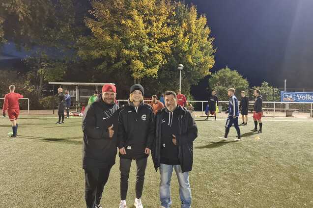 Drei Personen stehen auf einem Fu&szlig;ballplatz, umgeben von Spielern im Hintergrund unter Flutlicht.