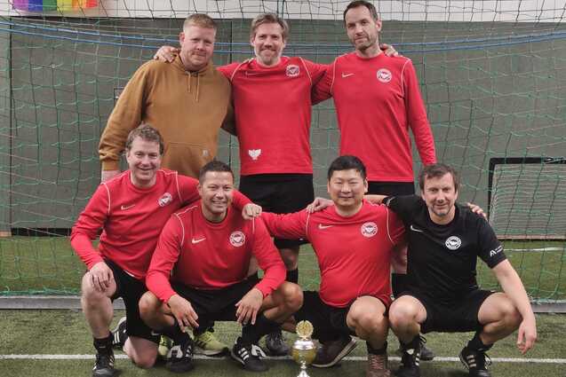 F&uuml;nf M&auml;nner in roten Trikots und zwei in schwarzem Trikot stehen l&auml;chelnd vor einem Fu&szlig;balltor mit einem Pokal.