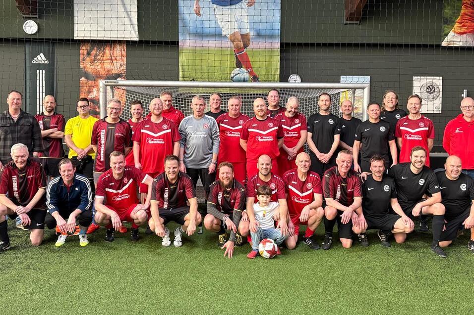 Gruppe von Erwachsenen in Fu&szlig;balltrikots posiert vor einem Tor auf einem Kunstrasenplatz.