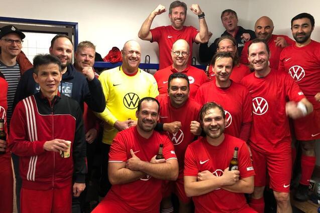 Mannschaft aus 15 M&auml;nnern in roten Fu&szlig;balltrikots posiert fr&ouml;hlich in einer Umkleidekabine mit Getr&auml;nken.