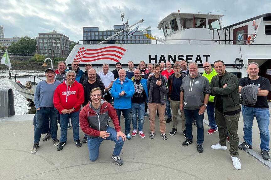 Gruppe von M&auml;nnern posiert l&auml;chelnd vor dem Boot "HANSEAT" an einem Hafen mit grauem Himmel.