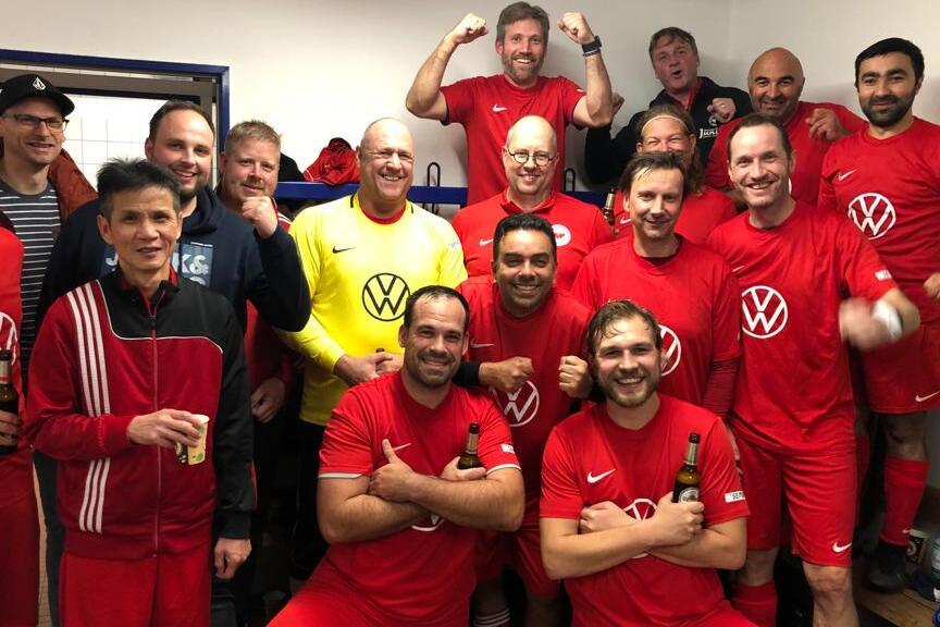 Mannschaft aus 15 M&auml;nnern in roten Fu&szlig;balltrikots posiert fr&ouml;hlich in einer Umkleidekabine mit Getr&auml;nken.