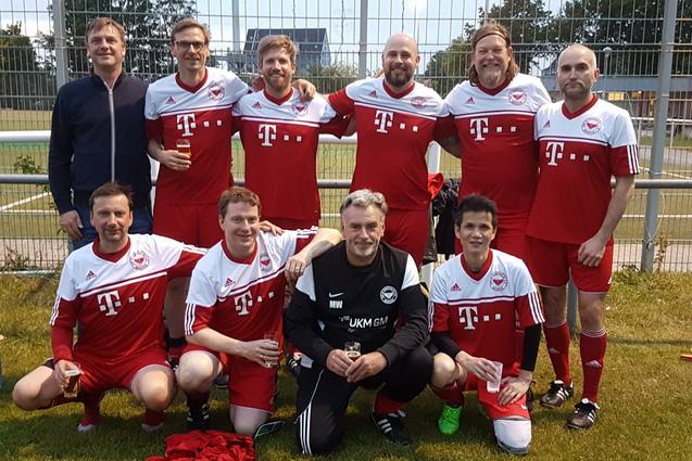 Mannschaftsfoto von zehn Fu&szlig;ballspielern in roten Trikots mit wei&szlig;em Logo, vor einem Fu&szlig;ballplatz bei D&auml;mmerung.