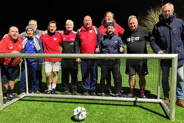 Gruppe von neun M&auml;nnern posiert l&auml;chelnd vor einem Fu&szlig;balltor auf einem Kunstrasenplatz in der Abendd&auml;mmerung.