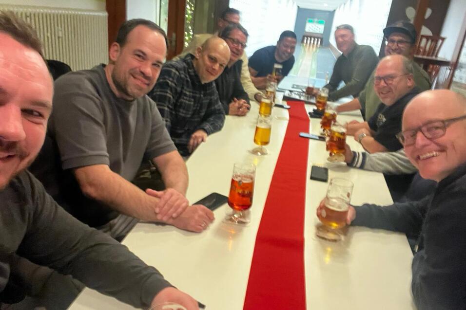Gruppenselfie von zehn M&auml;nnern an einem Tisch mit Bierkr&uuml;gen, auf einer Tischdecke mit rotem Streifen.