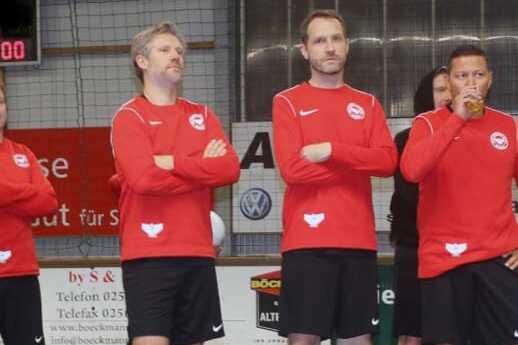 F&uuml;nf M&auml;nner in roten Fu&szlig;balltrikots stehen auf einem Sportfeld, einige mit verschr&auml;nkten Armen oder in Aktion.