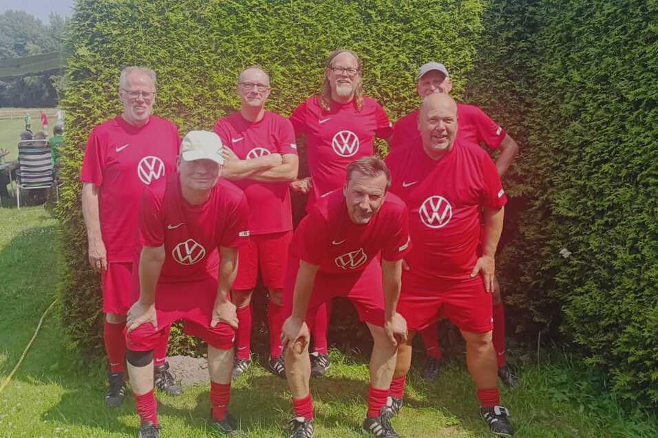 Sechs M&auml;nner in roten Trikots mit VW-Logo posieren auf einem Rasenfeld vor Hecken. Hintergrund mit Sitzen f&uuml;r Zuschauer.