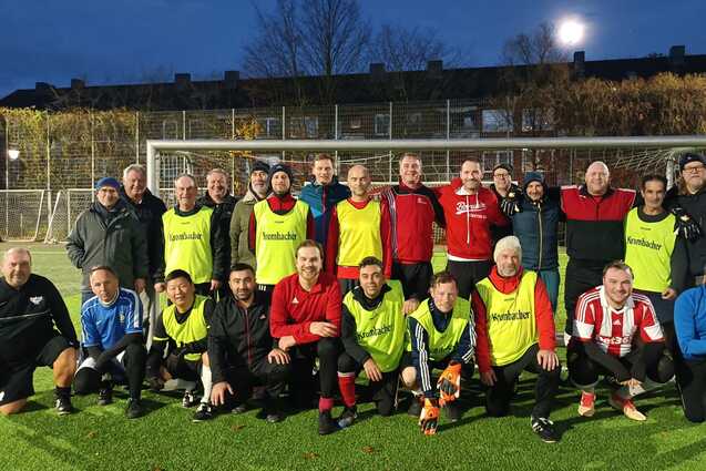 Gruppenfoto von Fu&szlig;ballspielern in Trikots und Trainingswesten auf einem Kunstrasenplatz bei Abendd&auml;mmerung.