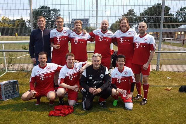 Mannschaftsfoto von zehn Fu&szlig;ballspielern in roten Trikots mit wei&szlig;em Logo, vor einem Fu&szlig;ballplatz bei D&auml;mmerung.