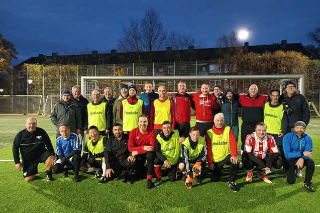 Gruppenfoto von Fu&szlig;ballspielern in Trikots und Trainingswesten auf einem Kunstrasenplatz bei Abendd&auml;mmerung.
