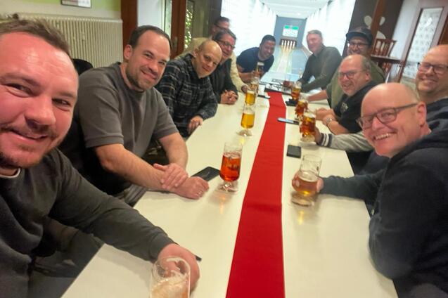Gruppenselfie von zehn M&auml;nnern an einem Tisch mit Bierkr&uuml;gen, auf einer Tischdecke mit rotem Streifen.