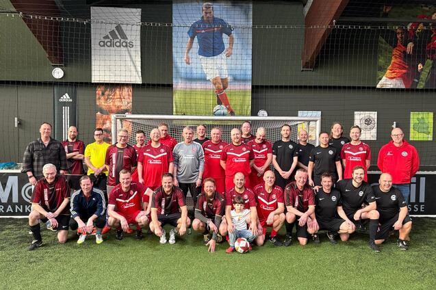 Gruppe von Erwachsenen in Fu&szlig;balltrikots posiert vor einem Tor auf einem Kunstrasenplatz.
