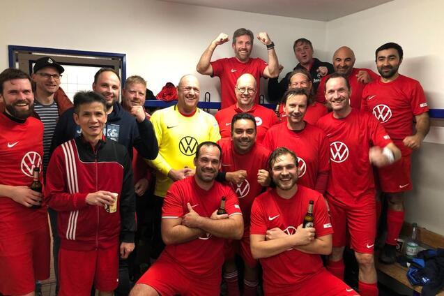 Mannschaft aus 15 M&auml;nnern in roten Fu&szlig;balltrikots posiert fr&ouml;hlich in einer Umkleidekabine mit Getr&auml;nken.