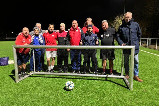 Gruppe von neun M&auml;nnern posiert l&auml;chelnd vor einem Fu&szlig;balltor auf einem Kunstrasenplatz in der Abendd&auml;mmerung.