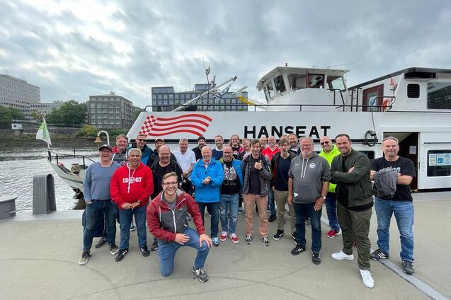 Gruppe von M&auml;nnern posiert l&auml;chelnd vor dem Boot "HANSEAT" an einem Hafen mit grauem Himmel.
