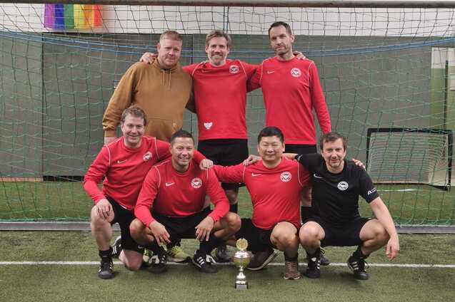 F&uuml;nf M&auml;nner in roten Trikots und zwei in schwarzem Trikot stehen l&auml;chelnd vor einem Fu&szlig;balltor mit einem Pokal.