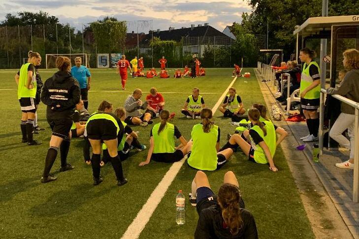 Mannschaftsbesprechung auf dem Fu&szlig;ballplatz mit Spielerinnen in gelben Trikots und einem Trainer im Abendlicht.