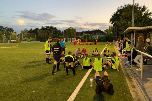 Mannschaftsbesprechung auf dem Fu&szlig;ballplatz mit Spielerinnen in gelben Trikots und einem Trainer im Abendlicht.