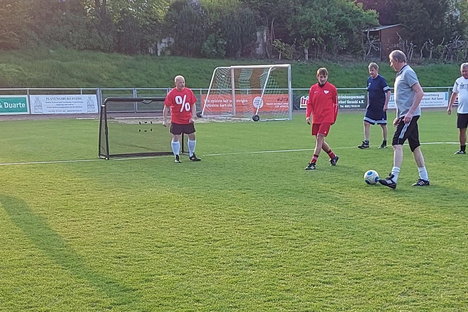 F&uuml;nf M&auml;nner in Sportkleidung &uuml;ben Fu&szlig;ball auf einem Rasenplatz mit einem kleinen Tor im Hintergrund.
