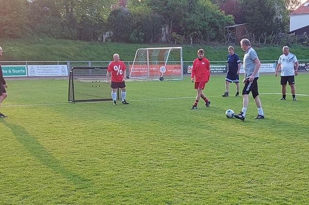 F&uuml;nf M&auml;nner in Sportkleidung &uuml;ben Fu&szlig;ball auf einem Rasenplatz mit einem kleinen Tor im Hintergrund.