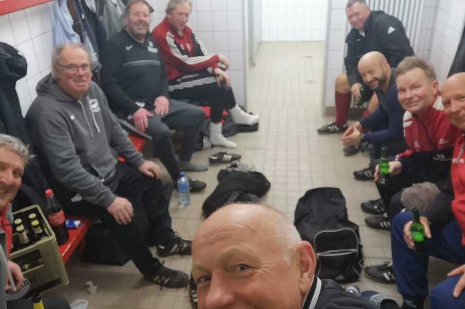 Gruppe von M&auml;nnern und einer Frau in Sportkleidung sitzt in einer Umkleidekabine, l&auml;chelnd und mit Getr&auml;nken.