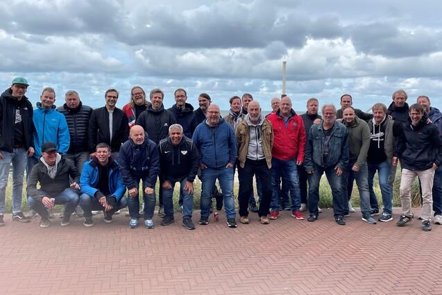 Gruppiert stehen 30 M&auml;nner auf einem Steinweg am Meer, mit Wolken und Wasser im Hintergrund.