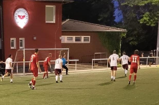 Fu&szlig;ballspiel bei D&auml;mmerung, Spieler in roter und wei&szlig;er Kleidung vor einem Vereinsgeb&auml;ude und Zuschauern am Rand.