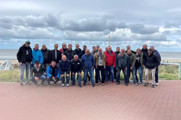 Gruppiert stehen 30 M&auml;nner auf einem Steinweg am Meer, mit Wolken und Wasser im Hintergrund.