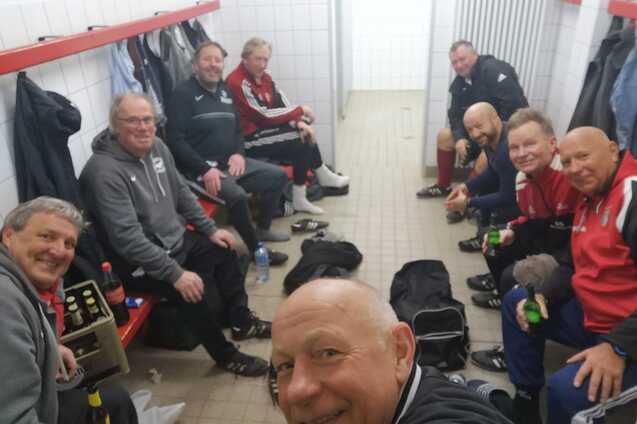 Gruppe von M&auml;nnern und einer Frau in Sportkleidung sitzt in einer Umkleidekabine, l&auml;chelnd und mit Getr&auml;nken.