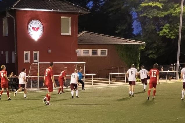 Fu&szlig;ballspiel bei D&auml;mmerung, Spieler in roter und wei&szlig;er Kleidung vor einem Vereinsgeb&auml;ude und Zuschauern am Rand.