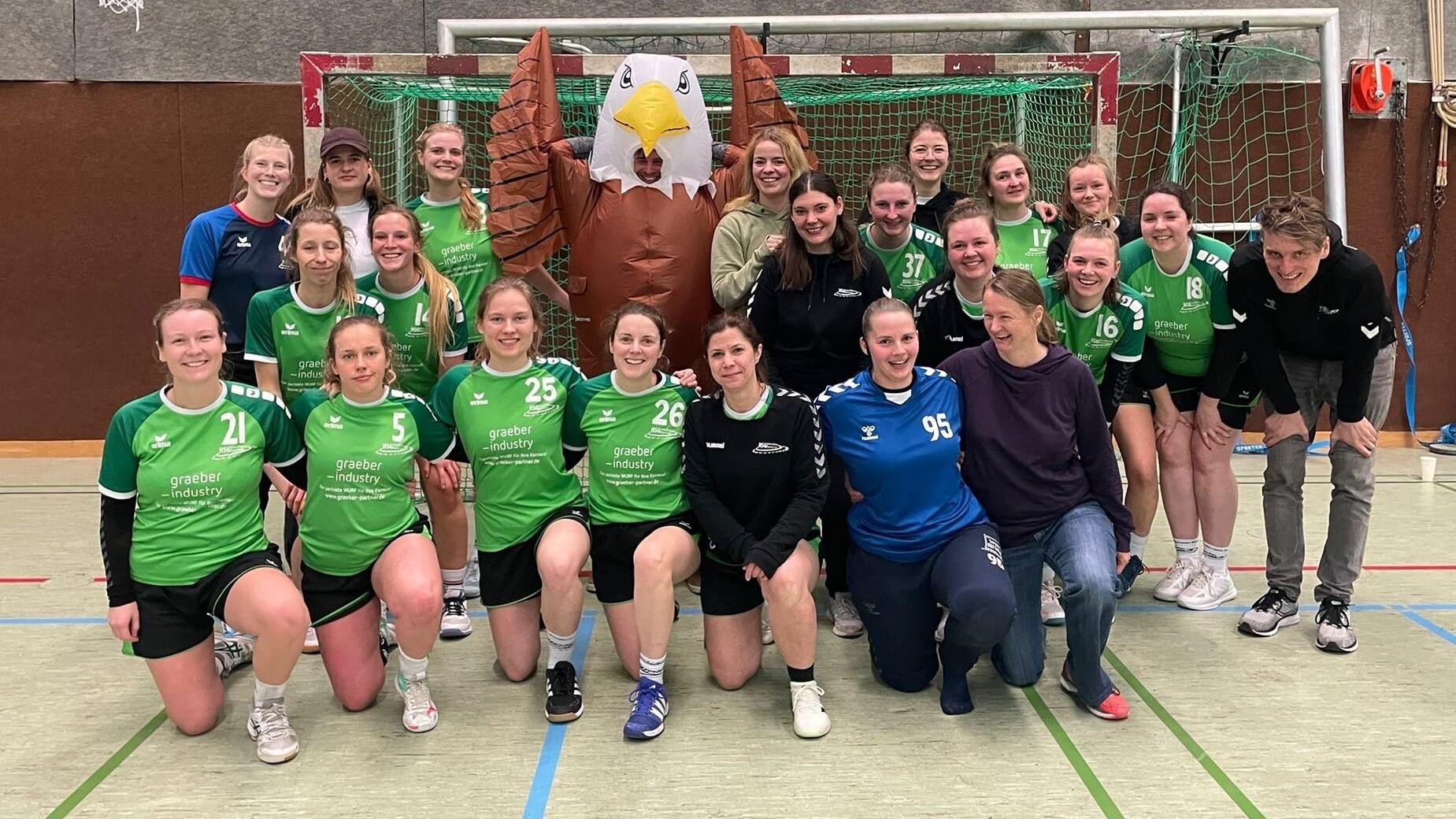 Gruppe von Handballspielerinnen in gr&uuml;nen Trikots posiert vor einem Tor mit einem gro&szlig;en Adler-Banner.