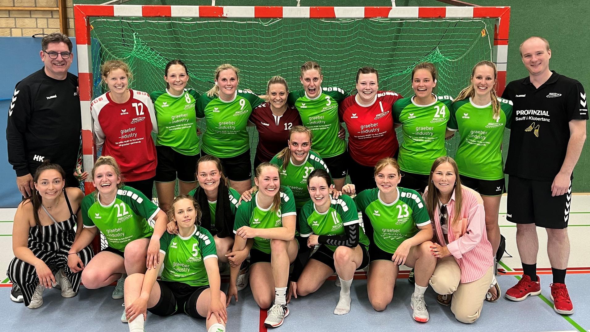 Gruppennfoto einer Handballmannschaft mit 14 Spielerinnen und zwei Trainern in Sportkleidung vor einem Tor.