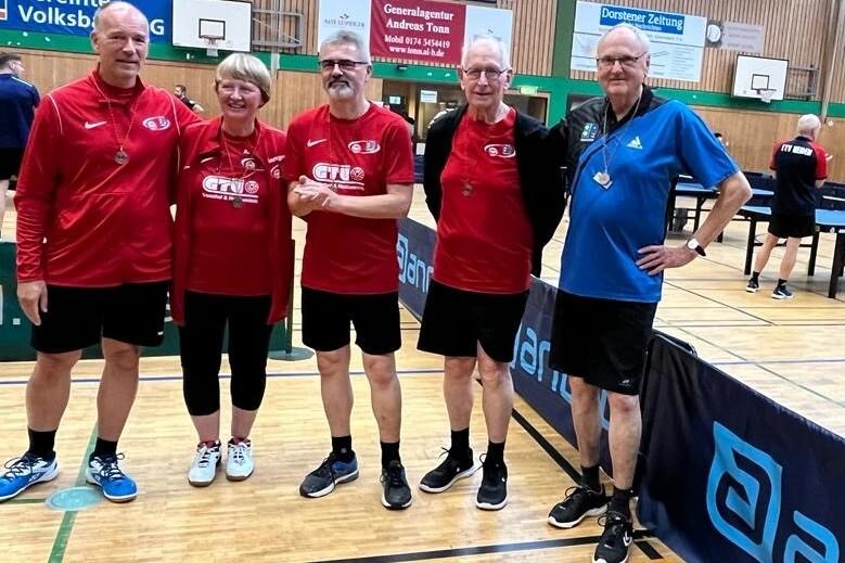F&uuml;nf Tischtennisspieler in roten und blauen Trikots posieren im Innenraum einer Sporthalle neben Tischtennisplatten.