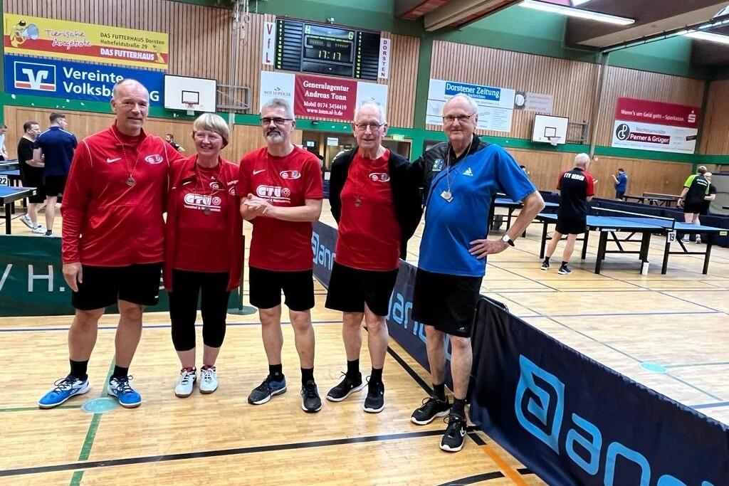 Fünf Tischtennisspieler in roten und blauen Trikots posieren im Innenraum einer Sporthalle neben Tischtennisplatten.