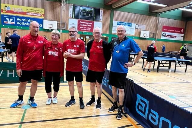 F&uuml;nf Tischtennisspieler in roten und blauen Trikots posieren im Innenraum einer Sporthalle neben Tischtennisplatten.