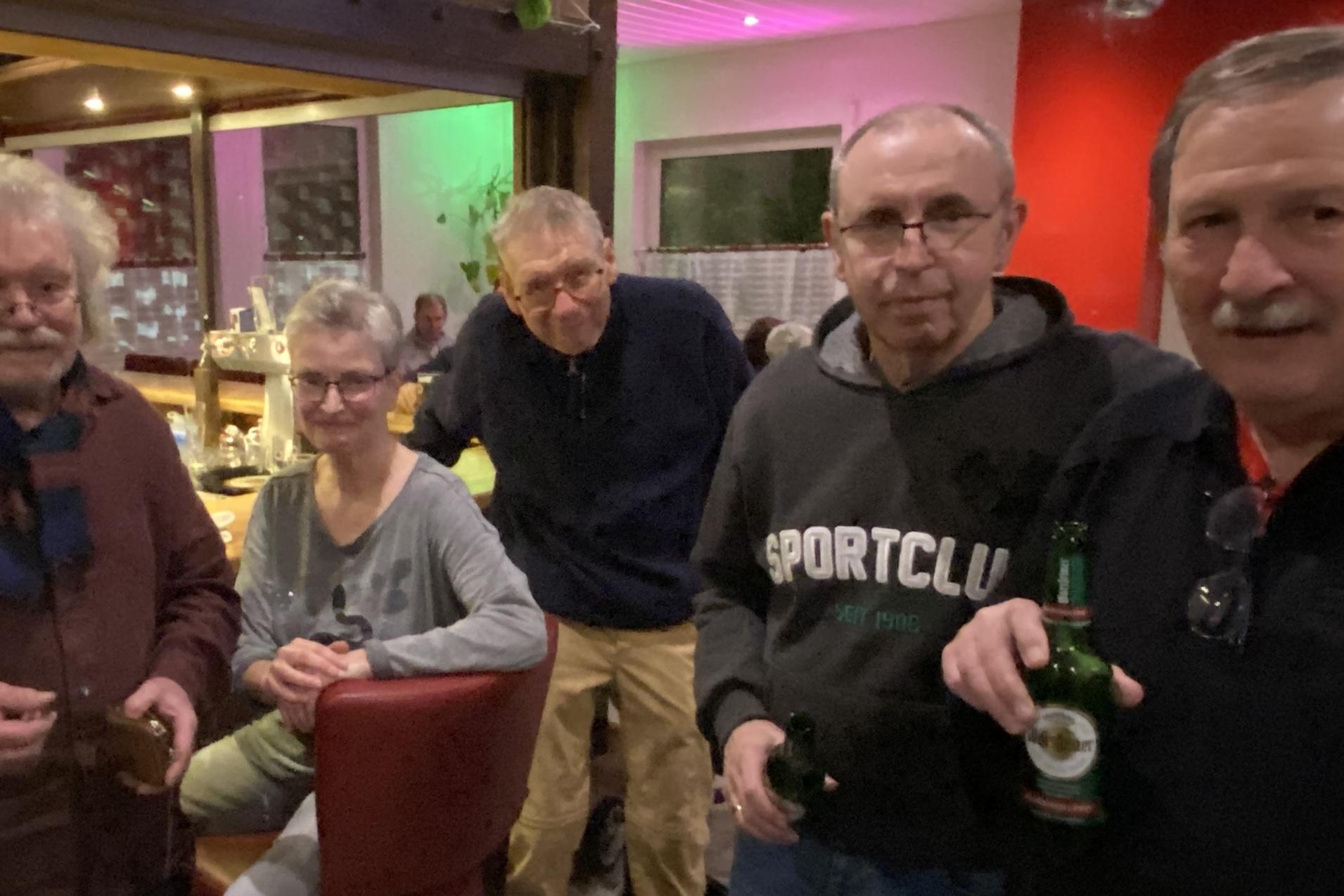 Gruppe von f&uuml;nf Personen steht in einer Bar, einige halten Getr&auml;nke, bunte Dekoration h&auml;ngt an der Decke.